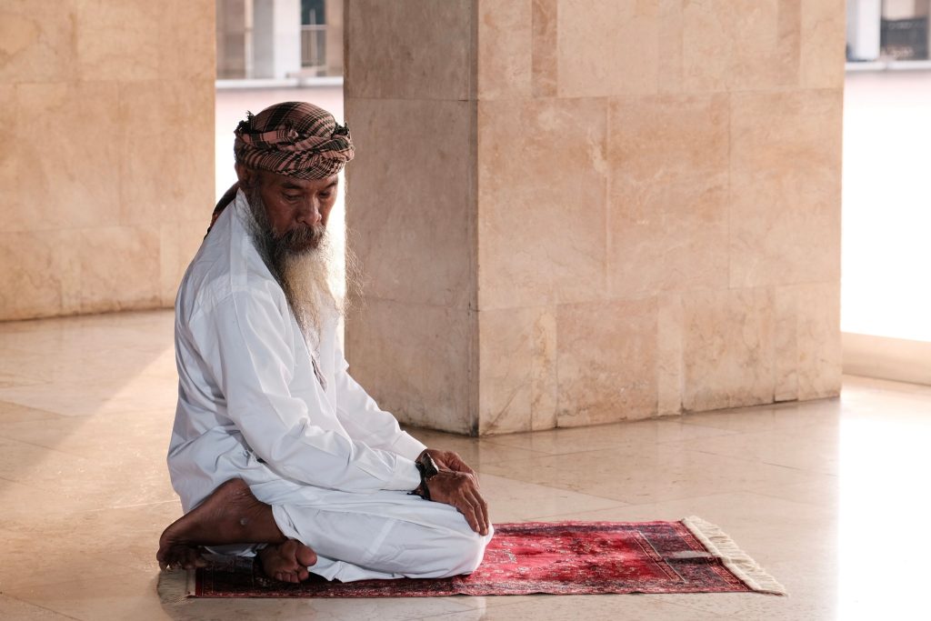 An elderly man in traditional attire praying inside a mosque in Jakarta, Indonesia.